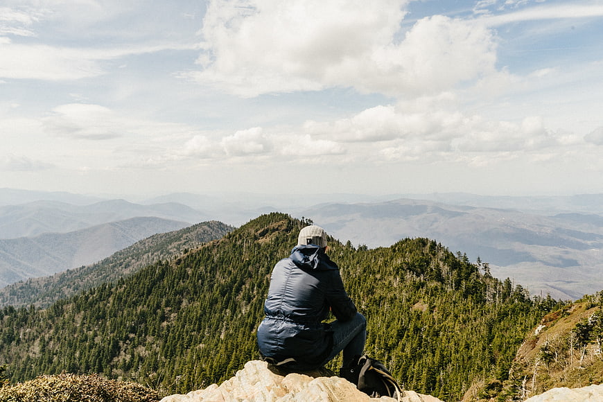 Man Sitting on Mountain Summit Overlooking Forested Peaks
