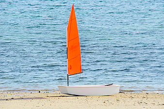 Small white sailboat with orange sail on sandy beach