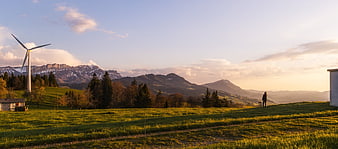 Wind Turbine In Alpine Pasture With Distant Snowy Peaks At Golden Hour