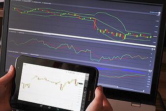 Hands holding Samsung tablet displaying DAX stock chart in front of laptop monitor