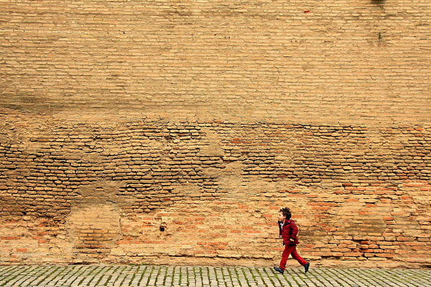 Child in red running past a massive textured ancient brick wall