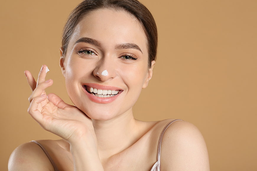 Happy woman applying face cream on beige background