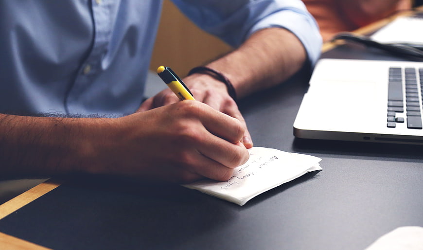 Man Writing Notes on Notepad at Desk with Laptop