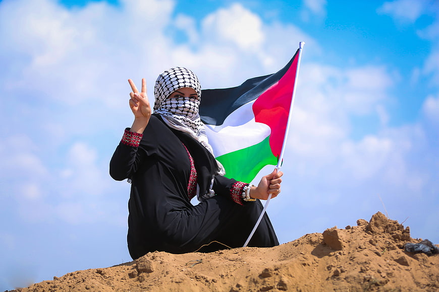 Woman in keffiyeh holding Palestinian flag on sandy hill