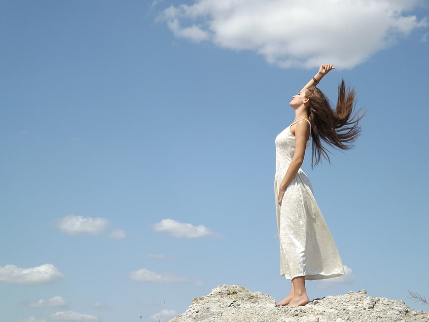 Woman in white dress standing on rocky cliff under blue sky