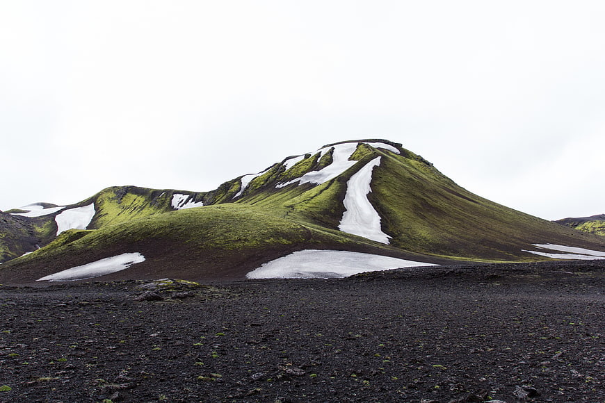 Green mossy mountain with snow patches on black volcanic sand