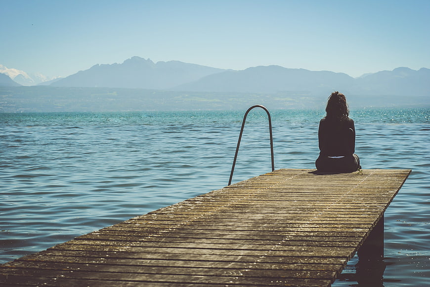 Woman sitting alone on wooden dock overlooking lake and mountains