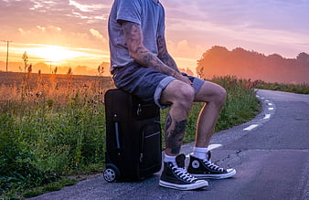 Tattooed Man Sitting on Suitcase by Road at Sunset
