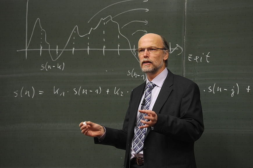 Senior male professor teaching physics in front of a green chalkboard