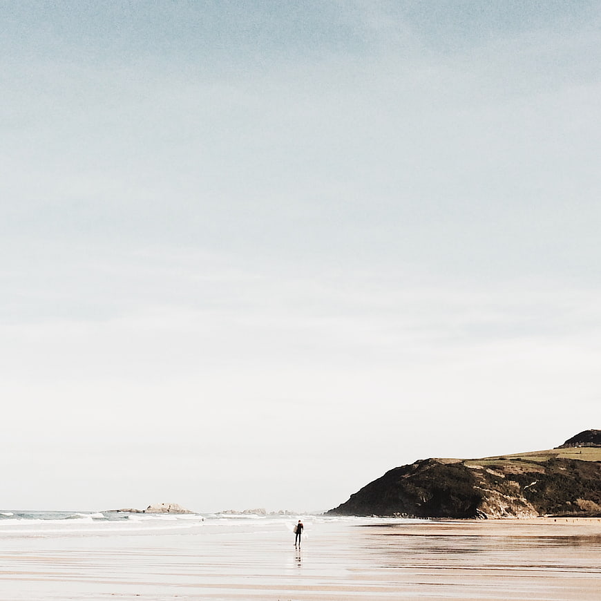 Lone Surfer Walking on Sandy Beach with Ocean and Hills
