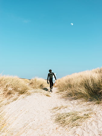Surfer walking through sand dunes with surfboard under blue sky