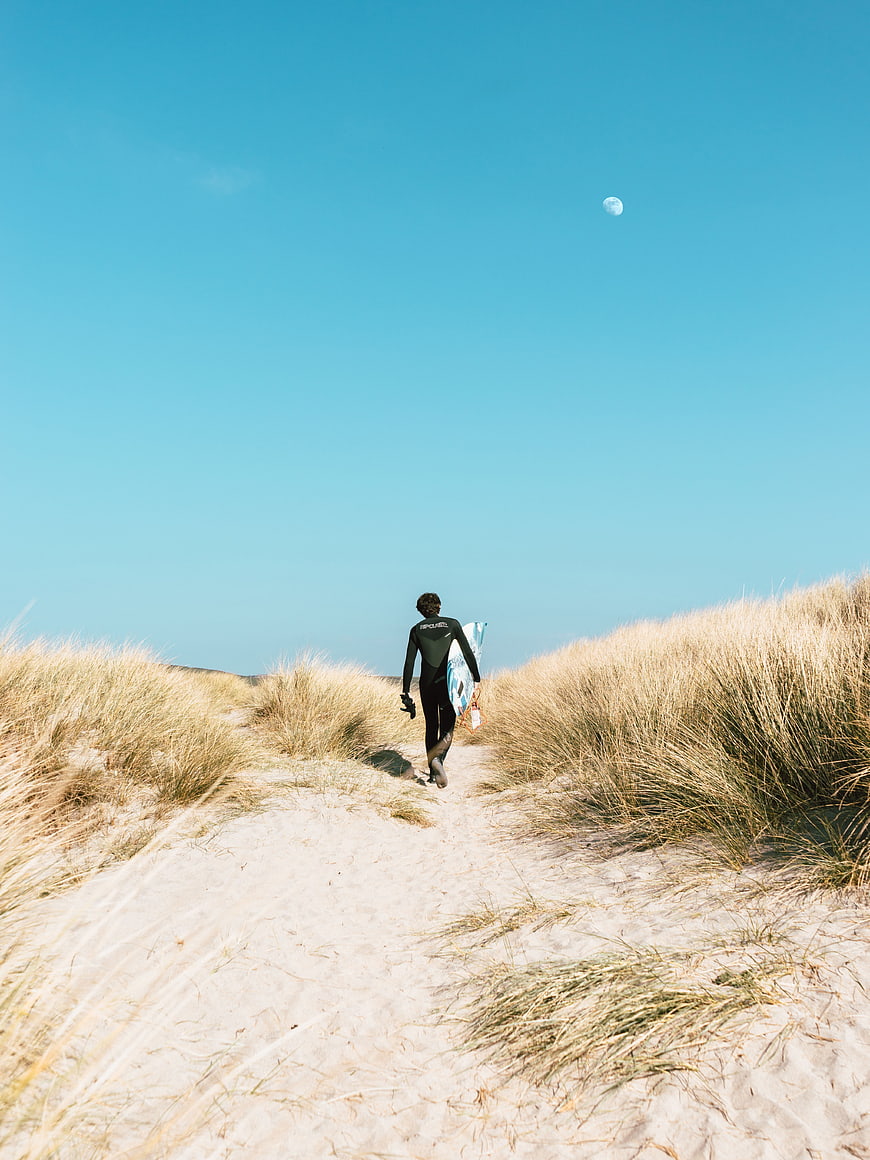 Surfer walking through sand dunes with surfboard under blue sky