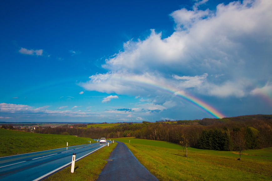 Rainbow over winding country road with car and green hills