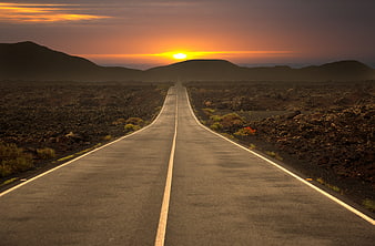 Straight asphalt road leading to sunset over volcanic landscape