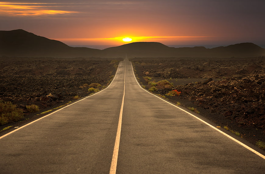 Straight asphalt road leading to sunset over volcanic landscape
