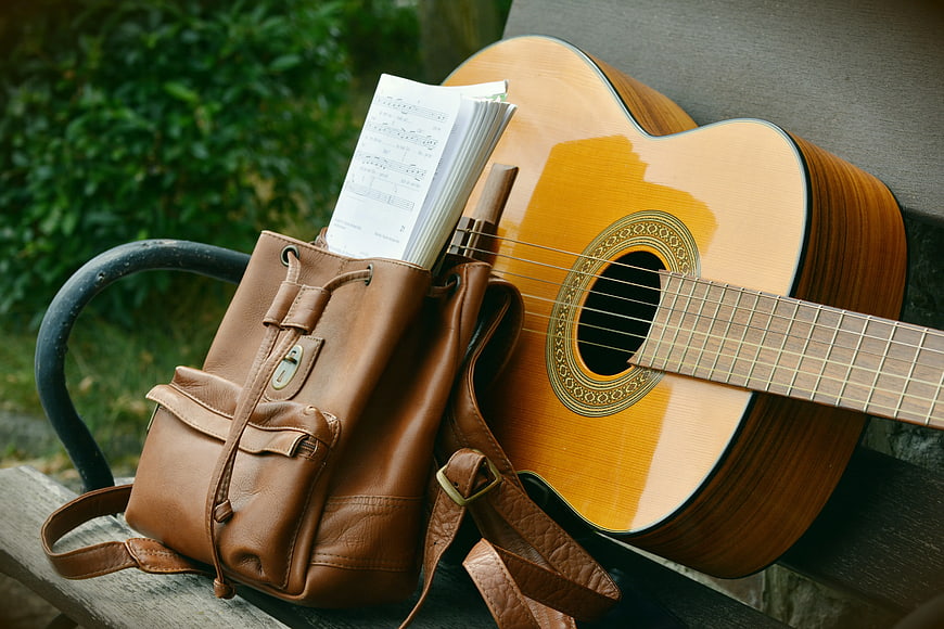 Acoustic Guitar and Leather Backpack with Sheet Music on Bench