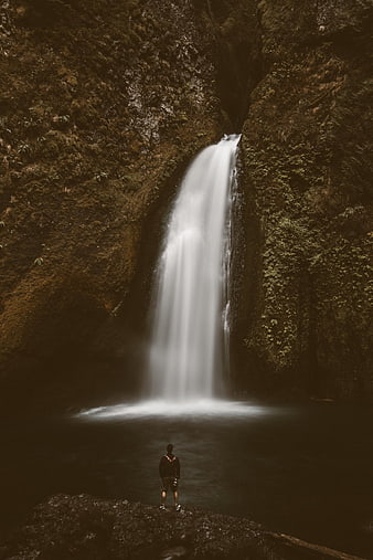 Lone figure standing before a long-exposure waterfall in a dark, mossy canyon