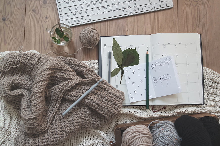 Flat lay of knitting supplies, planner, and yarn on wooden desk