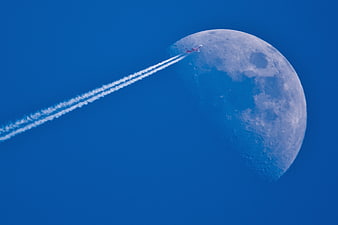 Airplane with contrails flying past a large gibbous moon in a blue sky
