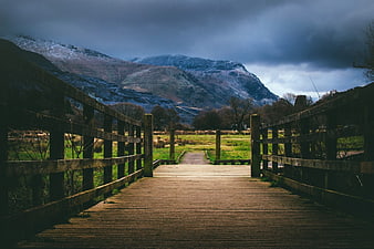 Wooden Bridge Leading to Snowy Mountains Under Dramatic Sky