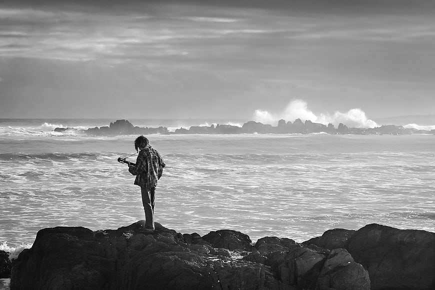 Lonely Man Playing Guitar On Rocky Coastline In Black And White