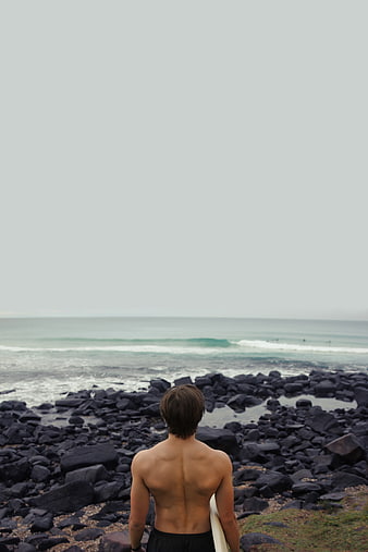 Surfer with surfboard looking out at ocean waves from rocky shore