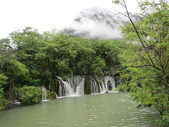Misty waterfall cascading into a serene green lake surrounded by lush forest