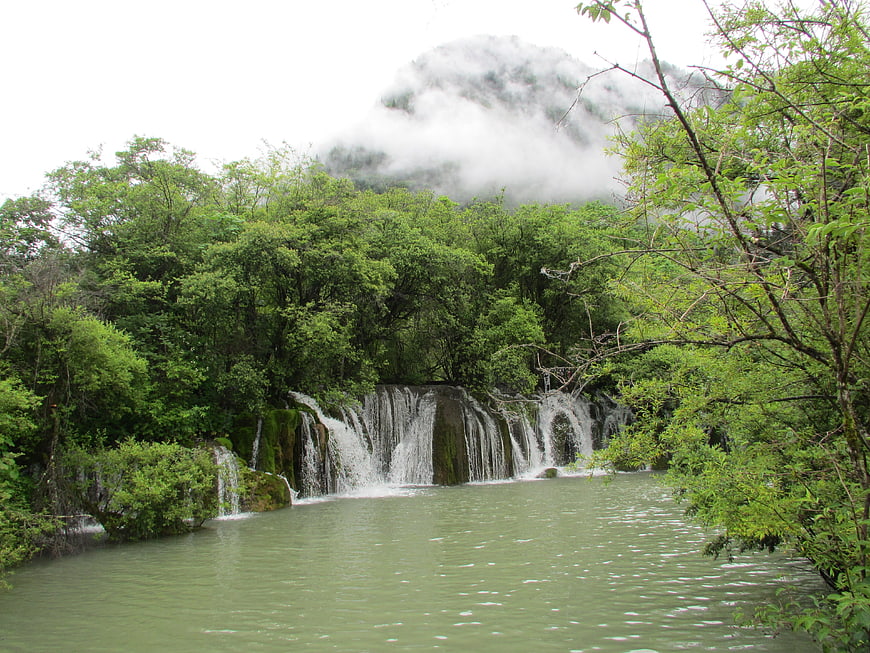 Misty waterfall cascading into a serene green lake surrounded by lush forest