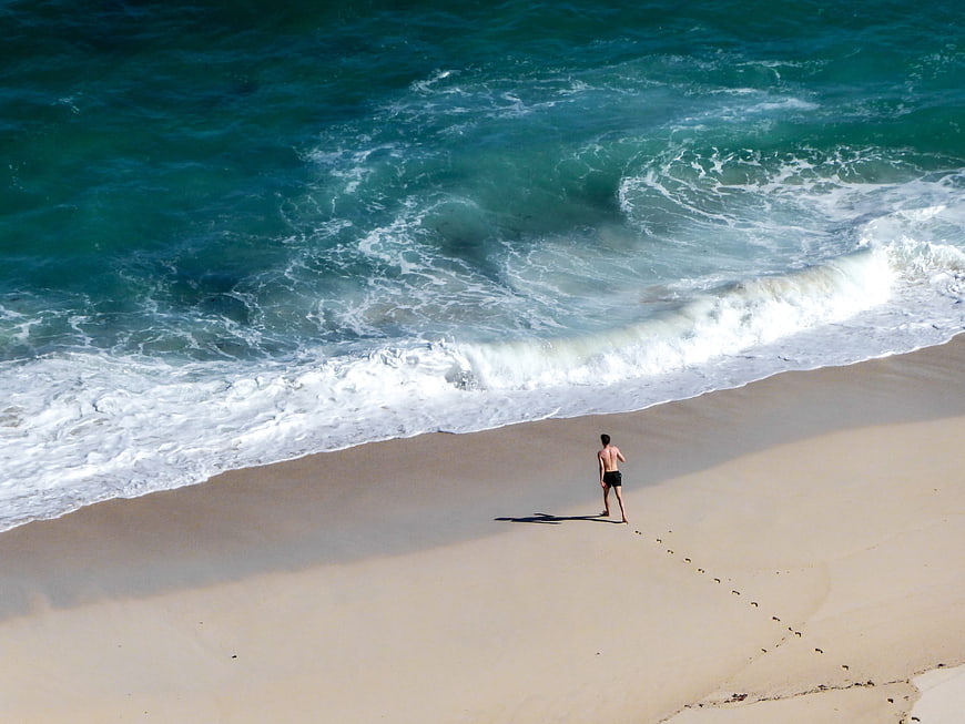 Aerial View of Man Walking on Sandy Beach Near Ocean Waves