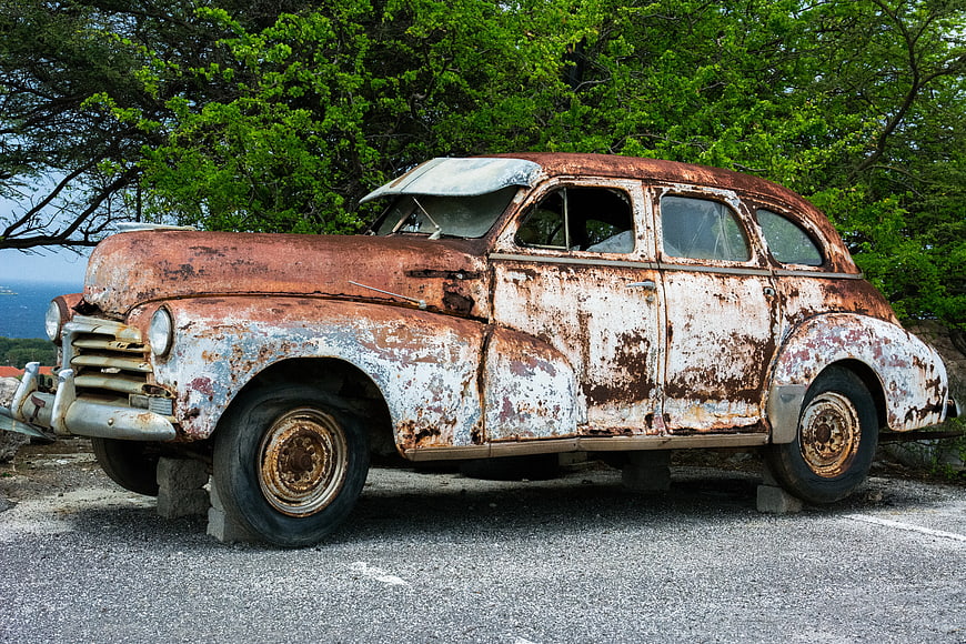 Rusty vintage car parked on asphalt near green trees