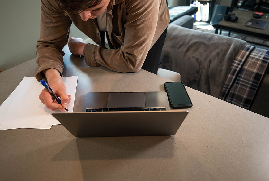 Man writing on paper at desk with laptop and smartphone