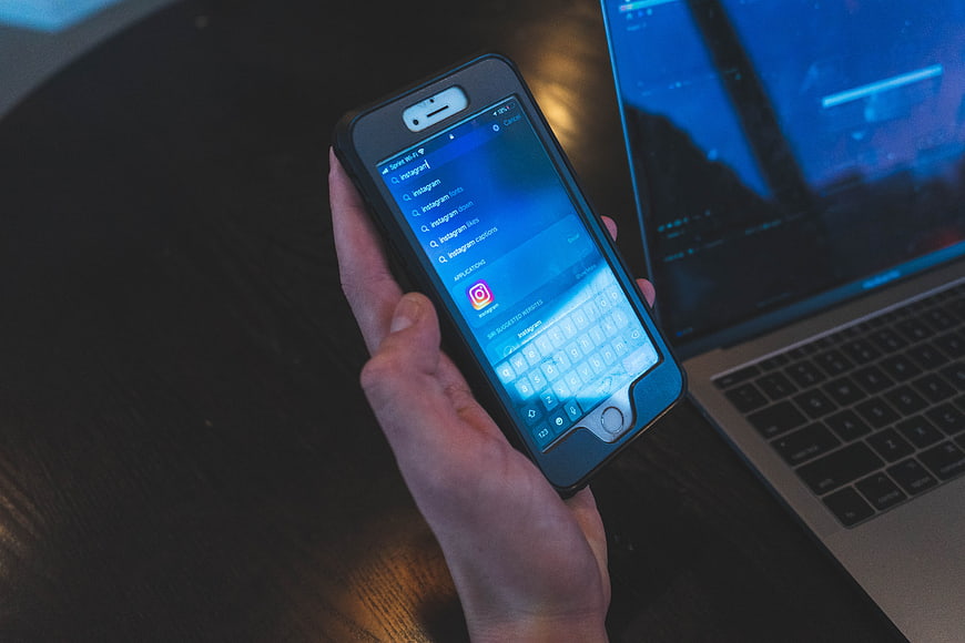 Hand holding smartphone searching for Instagram on laptop