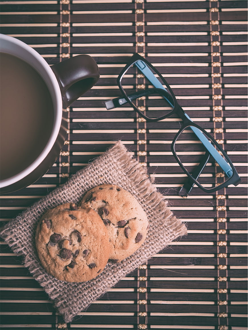 Flat lay of chocolate chip cookies, coffee mug, and glasses on bamboo mat