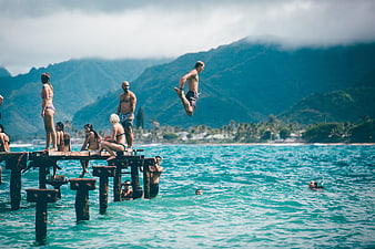 Man jumping off wooden pier into turquoise ocean with mountains