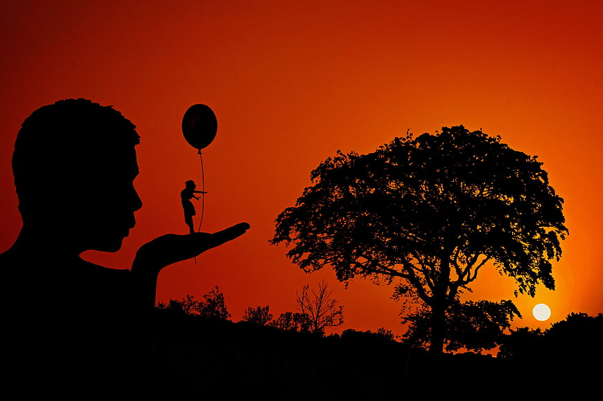 Silhouette of man holding tiny child with balloon at sunset