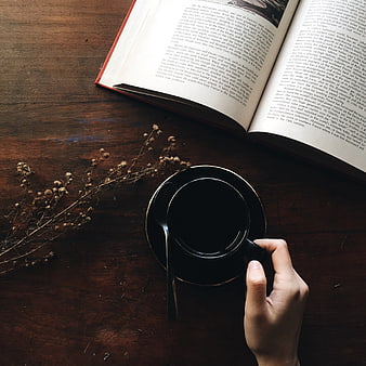 Hand holding black coffee cup on rustic wooden table with open book and dried flowers