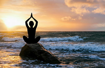 Silhouette of person meditating on rock at ocean sunset
