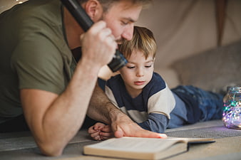Father and son reading with flashlight in cozy indoor tent