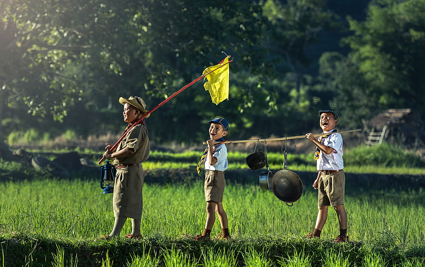 Three happy Thai scout boys hiking through green rice paddy field