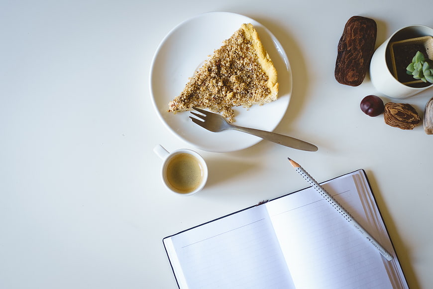 Flat lay of crumble cake, espresso, and open notebook on white desk