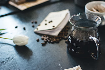 Pour-over coffee server with beans and white tulip on dark table