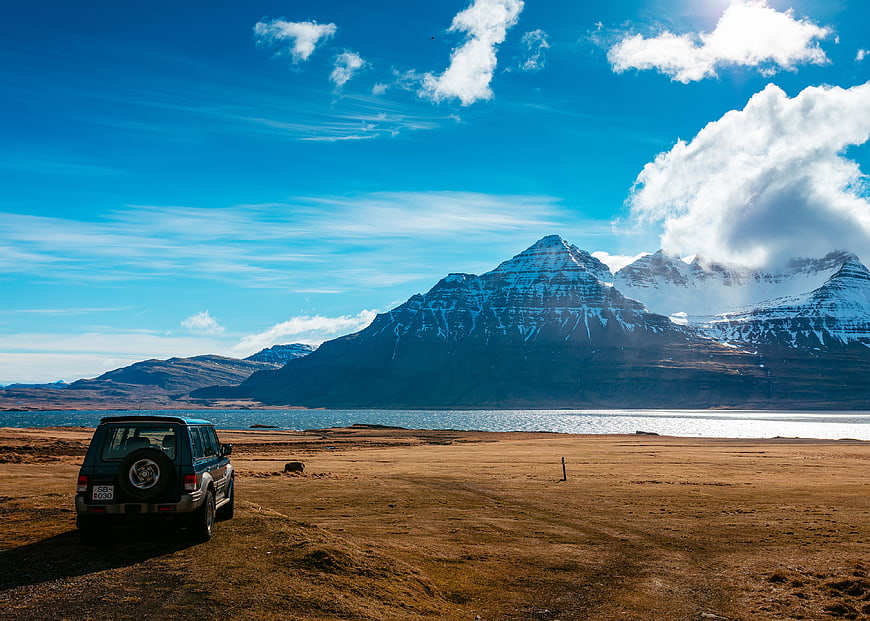 SUV parked on grassy shore facing snowy mountains and lake