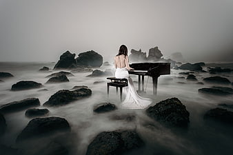 Woman in white dress playing grand piano on foggy rocky ocean shore