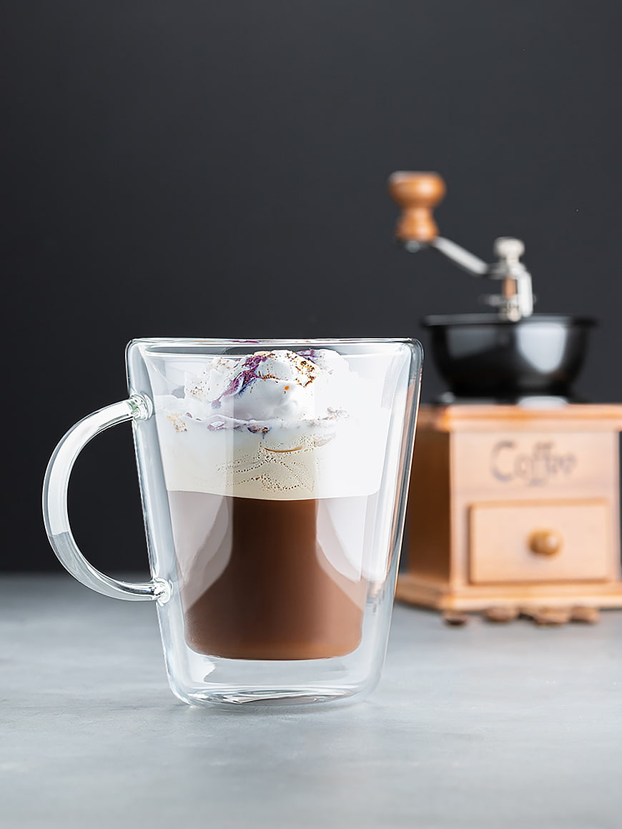 Iced coffee in double-walled glass mug with foam and coffee grinder background