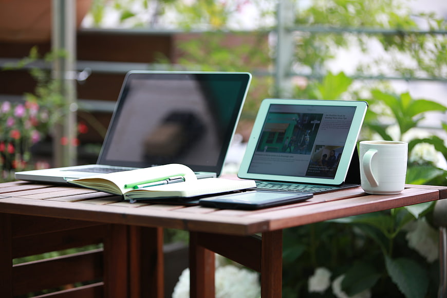 Outdoor workspace with laptop, tablet, notebook, and coffee on wooden table