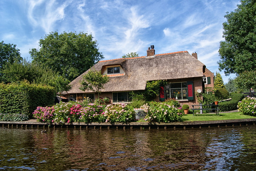 Traditional Dutch Thatched Cottage With Vibrant Garden By Canal