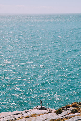 Lone figure standing on rocky cliff overlooking turquoise ocean