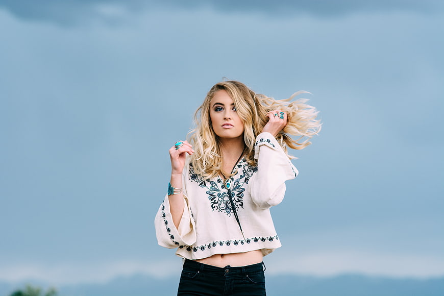 Blonde woman in embroidered top posing against blue sky