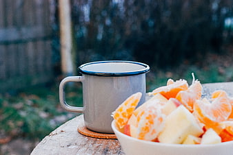 Enamel mug and fresh fruit bowl on rustic wooden stump outdoors