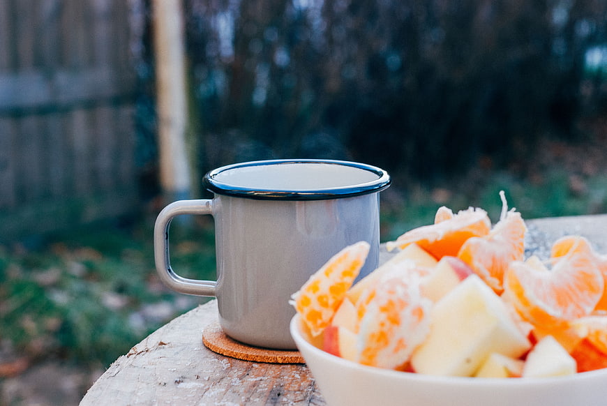 Enamel mug and fresh fruit bowl on rustic wooden stump outdoors